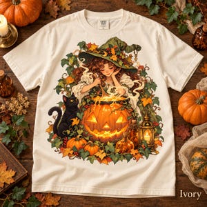 May include: An ivory t-shirt featuring a graphic illustration of a person wearing a green witch hat, stirring a glowing jack-o'-lantern cauldron. A black cat sits beside the pumpkin, surrounded by autumn leaves, vines, and a lantern.