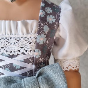Pode incluir: Close-up de um vestido tradicional de boneca. O vestido apresenta um padrão floral cinzento no corpete e nas mangas, com renda branca e um laço azul. Um pendente em forma de coração pende de uma fita.
