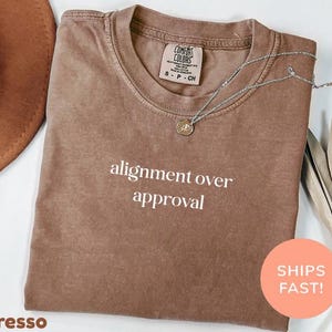 May include: A brown short-sleeved t-shirt with a crew neckline featuring the white text alignment over approval printed on the chest. The shirt has a Comfort Colors brand label at the inner neck and is displayed with a silver chain necklace.