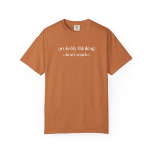 Pode incluir: Camiseta cor de ferrugem com a frase "probably thinking about snacks" impressa em branco. A camiseta de manga curta tem gola redonda e é feita de um tecido macio.