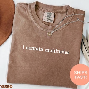 May include: Espresso-colored t-shirt with the phrase "i contain multitudes" in white. The shirt has a crew neck and a small tag on the upper back. A silver necklace with a pendant is draped across the shirt. A brown hat and dried leaves are in the background.