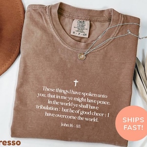 Könnte beinhalten: Ein braunes T-Shirt mit kurzen Ärmeln, einem weißen Kreuz und dem Text: These things I have spoken unto you, that in me ye might have peace. In the world ye shall have tribulation: but be of good cheer; I have overcome the world. John 16:33.