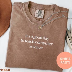 May include: Espresso-colored t-shirt with the text "it's a good day to teach computer science" in white. A silver necklace with a pendant rests on the shirt. A brown hat and dried leaves are in the background.