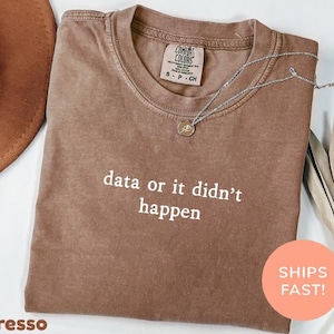 Puede incluir: Camiseta color espresso con el texto blanco "data or it didn't happen". La camiseta tiene cuello redondo y mangas cortas. Un collar plateado con un colgante está drapeado sobre la camiseta. La camiseta está doblada sobre una superficie blanca.