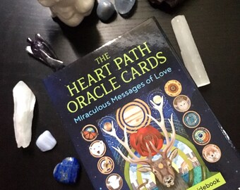 以下が含まれることがあります： 「The Heart Path Oracle Cards: Miraculous Messages of Love」というタイトルのオラクルカードデッキ。53枚のカードとガイドブック付き。表紙には大きな角を持つ鹿と赤いバラが描かれています。様々なクリスタルと小さな白い置物がデッキを囲んでいます。