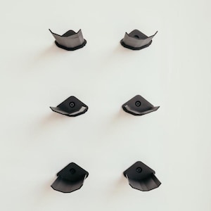 May include: Six dark gray corner protectors are mounted on a white wall. Each protector has a triangular shape with a curved edge and a circular fastener in the center. The protectors are evenly spaced and arranged in a grid pattern.