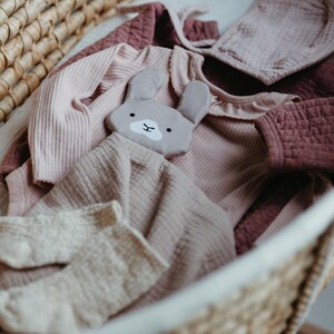 May include: A woven basket filled with baby clothes in shades of pink and mauve. Items include a ribbed long-sleeved onesie, a bonnet, a ruffled collar top, and a stuffed bunny toy. The basket is lined with a white fabric.