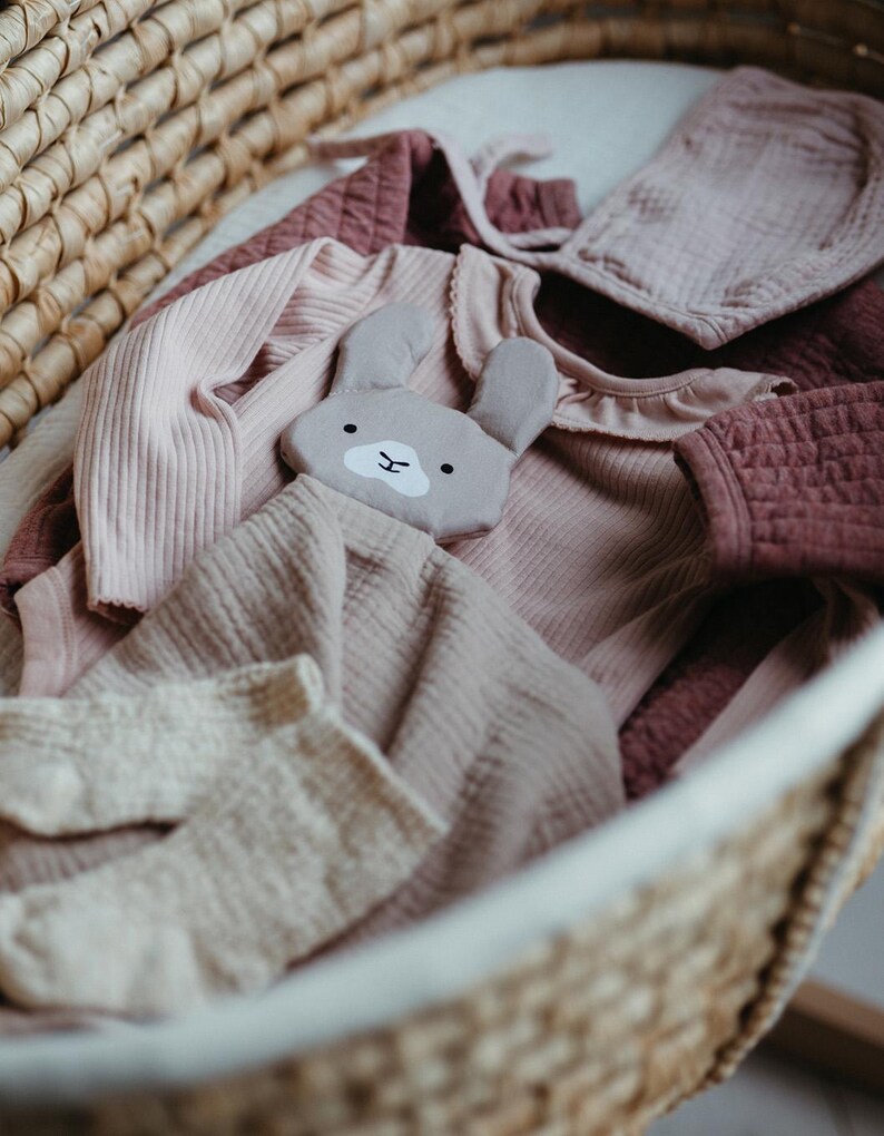 May include: A woven basket filled with baby clothes in shades of pink and mauve. Items include a ribbed long-sleeved onesie, a bonnet, a ruffled collar top, and a stuffed bunny toy. The basket is lined with a white fabric.