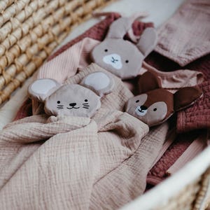 May include: A woven basket holds a collection of baby items. There are three plush animal head security blankets: a gray mouse, a gray bunny, and a brown deer. The blankets are on top of soft, pink and mauve blankets.