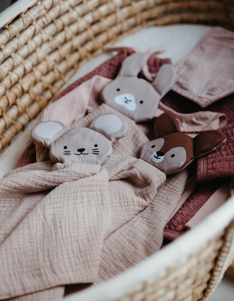 May include: A woven basket holds a collection of baby items. There are three plush animal head security blankets: a gray mouse, a gray bunny, and a brown deer. The blankets are on top of soft, pink and mauve blankets.
