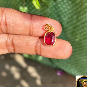May include: A small oval-shaped pendant featuring a vibrant, deep red gemstone set in a polished yellow gold frame. The pendant includes a simple gold loop at the top for attachment, showcasing a faceted surface that reflects light across the stone.