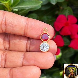 May include: A gold-toned pendant featuring two circular stones arranged vertically. The top stone is a vibrant red, while the bottom stone is clear and faceted. A small gold loop is attached to the top for hanging, creating a simple jewelry piece.