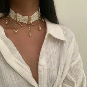 May include: A white beaded choker necklace with gold-colored accents and dangling pearl pendants. The necklace is worn over a white collared shirt. The choker has a rectangular gold-colored clasp.