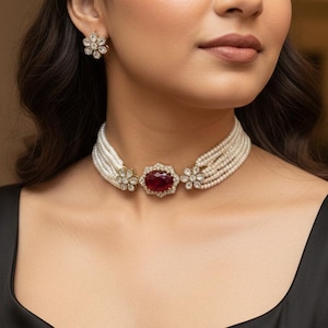 May include: A pearl choker necklace with a central ruby-colored gemstone and floral accents. Matching floral stud earrings are also visible. The jewelry is set against a black top, creating a contrast that highlights the necklace's design.