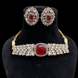 Peut inclure: Un ensemble de bijoux argentés comprenant un collier ras du cou et des boucles d'oreilles assorties. Chaque pièce comporte une grosse pierre centrale rouge rubis carrée entourée de petites pierres claires. L'ensemble est présenté sur un mannequin noir.