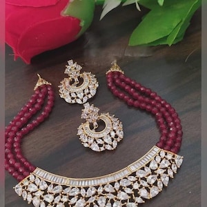 May include: A three-strand burgundy beaded necklace and matching earrings set. The necklace features a gold-toned base with clear, teardrop-shaped and baguette-cut crystals. The earrings have a similar design. The jewelry set is displayed on a dark wooden surface.