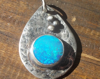 May include: A silver pendant with a teardrop shape and a textured surface. The pendant features a vibrant, round, blue opal stone set in a silver bezel. Three small silver beads are placed near the top.