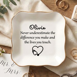 以下が含まれることがあります： 「Olivia」の名前と心温まるメッセージが刻まれた、金色の縁取りが施された白いセラミック製の小物入れ。聴診器のハートのグラフィックが含まれています。結婚指輪と花のアクセントが皿を囲み、心のこもった贈り物となっています。