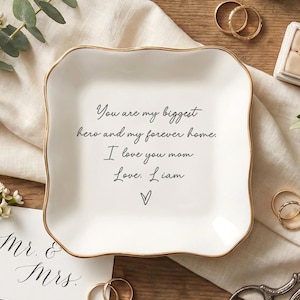 May include: A white ceramic trinket dish with a gold rim, inscribed with a heartfelt message. The dish is surrounded by wedding rings, a ring box, and floral accents, suggesting a wedding or anniversary theme.
