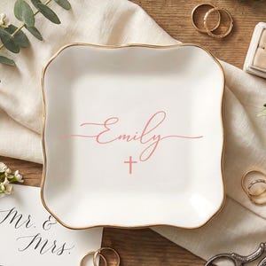 May include: A white ceramic ring dish with a gold rim, featuring the name "Emily" in pink script and a small cross. The dish is surrounded by wedding rings, greenery, and a "Mr. & Mrs." card.