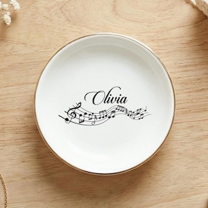 以下が含まれることがあります： 金色の縁取りのある白いセラミック製の小物入れ。名前「Olivia」と音符が黒い筆記体で描かれています。 金色のブレスレット、パールのイヤリング、レースが木製の表面に置かれています。