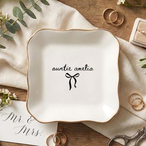 May include: A white ceramic trinket dish with a gold rim, featuring the text "auntie amelia" and a black bow design. The dish is surrounded by wedding rings, a ring box, and floral accents, suggesting a wedding or gift theme.