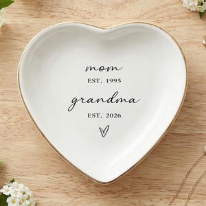 Peut inclure: Un vide-poches en céramique blanc en forme de cœur avec un bord doré. Le plat porte les inscriptions "maman EST. 1995" et "grand-mère EST. 2026". Le plat est entouré de brins de fleurs blanches et de colliers dorés.