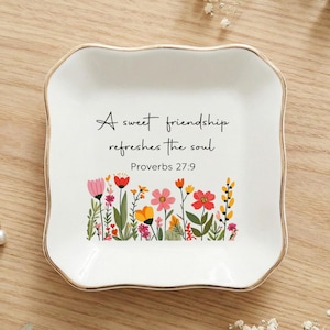 Op de afbeelding: Wit keramisch sieradenbakje met een gouden rand, met de quote "Een zoete vriendschap verfrist de ziel" en een bloemenontwerp. Inclusief een gouden ring met een parel en kleine zilveren oorbellen.
