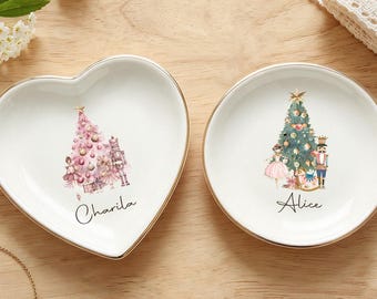 Puede incluir: Dos platos de cerámica blanca para joyas con bordes dorados. Uno tiene forma de corazón con un árbol de Navidad rosa, un cascanueces y el nombre Charila. El otro es circular con un árbol de Navidad verde, un cascanueces y el nombre Alice.