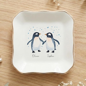 以下が含まれることがあります： 金色の縁取りが付いた白いセラミック製の小物入れ。手をつないでいる2羽のペンギンの水彩画イラストが描かれています。「Oliver」と「Sophie」の名前が下に書かれています。皿は、スカラップエッジの正方形です。