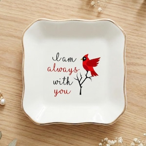 May include: White ceramic trinket dish with a gold rim, featuring a red cardinal perched on a branch and the text "I am always with you." A ring and earrings are also visible.