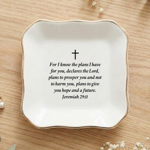 Puede incluir: Plato de cerámica blanca para baratijas con borde dorado, con una cruz negra y el versículo bíblico Jeremías 29:11. Un anillo dorado con una perla y dos pares de pendientes están junto al plato. El plato está sobre una superficie de madera.