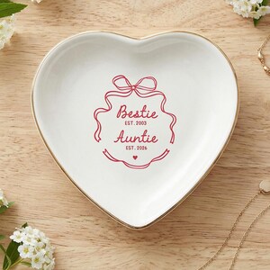 May include: A white heart-shaped ceramic trinket dish with a gold-toned rim. The center features a red graphic of a ribbon frame with the text "Bestie EST. 2003" and "Auntie EST. 2026" printed in a script and sans-serif font, with a small heart below.