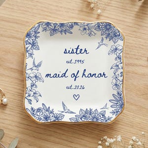 May include: A white ceramic trinket dish with a blue floral design and gold trim. The dish is inscribed with the words "sister est. 1995 maid of honor est. 2026" and a heart symbol. A ring and earrings are on the wooden surface.