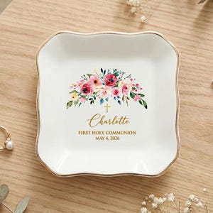 May include: A white ceramic trinket dish with a gold rim, featuring a floral design in pink, red, and yellow. The dish is personalized with the name "Charlotte", a cross, and the text "First Holy Communion, May 4, 2026".