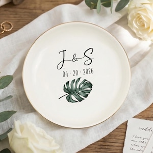 Puede incluir: Un plato de cerámica blanco con borde dorado, con las iniciales "J&S", la fecha "04.20.2026" y un diseño de hoja de monstera verde. El plato está rodeado de decoraciones temáticas de boda, incluyendo anillos y flores.