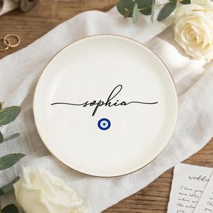 May include: A white ceramic plate with a gold rim, featuring the name "sophia" in elegant black script. Below the name is a blue and white evil eye symbol. The plate is surrounded by wedding-themed decorations.