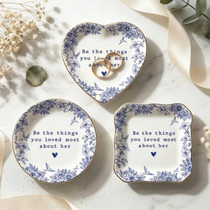 Op de afbeelding: Drie decoratieve keramische ringschalen in hart-, ronde en vierkante vormen. Elke schaal heeft een blauw bloemmotief met gouden rand en de tekst "Be the things you loved most about her".