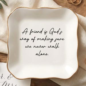 May include: A white ceramic ring dish with a gold rim, inscribed with the quote "A friend is God's way of making sure we never walk alone." The image includes wedding rings, a ring box, and floral accents.