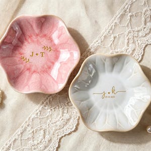 May include: Two scalloped, flower-shaped trinket dishes. One dish is pink with a marbled finish and gold lettering reading J + T with leaf accents. The other is white with a glossy finish and gold lettering reading j + k and 05.18.2025.