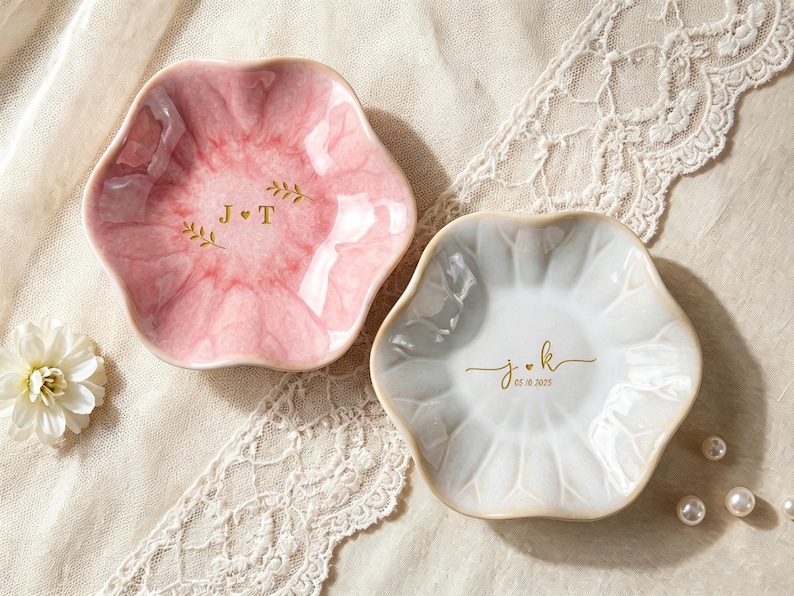 May include: Two scalloped, flower-shaped trinket dishes. One dish is pink with a marbled finish and gold lettering reading J + T with leaf accents. The other is white with a glossy finish and gold lettering reading j + k and 05.18.2025.