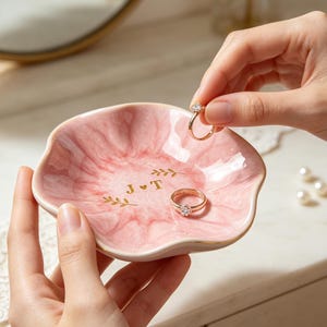 May include: A scalloped, pink marble-patterned ceramic ring dish featuring gold-colored text J heart T and small leaf sprigs in the center. The dish has a gold-painted rim and holds a gold ring with a clear stone, while another ring rests inside.