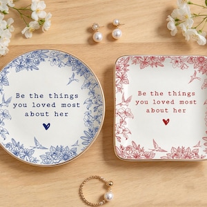 Może przedstawiać: Okrągła ceramiczna miseczka i kwadratowa ceramiczna miseczka, obie ze złotym brzegiem, kwiatowymi obw&oacute;dkami i napisem "Be the things you loved most about her" oraz małą ikoną serca. Okrągła miseczka ma niebieskie detale, a kwadratowa czerwone.