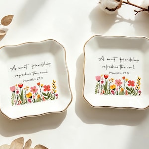 May include: Two square white ceramic trinket dishes with scalloped edges and gold-toned rims. Each dish features a colorful floral illustration at the bottom and the black text: A sweet friendship refreshes the soul. Proverbs 27:9.