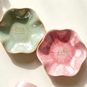May include: Two flower-shaped ceramic trinket dishes with a glossy finish. One dish is sage green with gold lettering k heart f and 07.02.2025. The other is dusty rose with gold lettering j heart k and 05.10.2025. Both have scalloped edges.