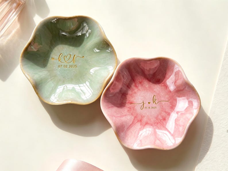 May include: Two flower-shaped ceramic trinket dishes with a glossy finish. One dish is sage green with gold lettering k heart f and 07.02.2025. The other is dusty rose with gold lettering j heart k and 05.10.2025. Both have scalloped edges.