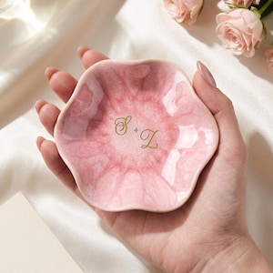 May include: A small, flower-shaped trinket dish with a glossy, marbled pink finish. The center of the dish features the gold-colored script letters S + Z. The dish has a wavy, scalloped edge and a smooth, reflective surface texture throughout.