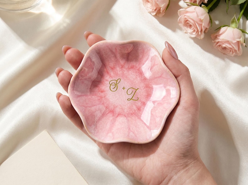 May include: A small, flower-shaped trinket dish with a glossy, marbled pink finish. The center of the dish features the gold-colored script letters S + Z. The dish has a wavy, scalloped edge and a smooth, reflective surface texture throughout.