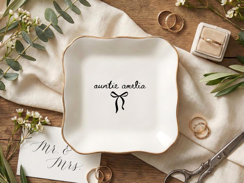 May include: A white ceramic trinket dish with a gold rim, featuring the text "auntie amelia" and a black bow design. The dish is surrounded by wedding rings, a ring box, and floral accents, suggesting a wedding or gift theme.