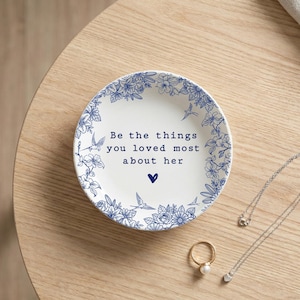 Może przedstawiać: Okrągła biała ceramiczna miseczka z niebieską kwiatową obw&oacute;dką z małymi ptakami oraz napisem "Be the things you loved most about her" wydrukowanym na środku nad małą niebieską ikoną serca, w towarzystwie złotego pierścionka i srebrnego naszyjnika.
